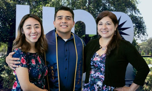 A smiling graduate wearing his Navy graduation sash poses with family in front of large metal letters spelling UCR.